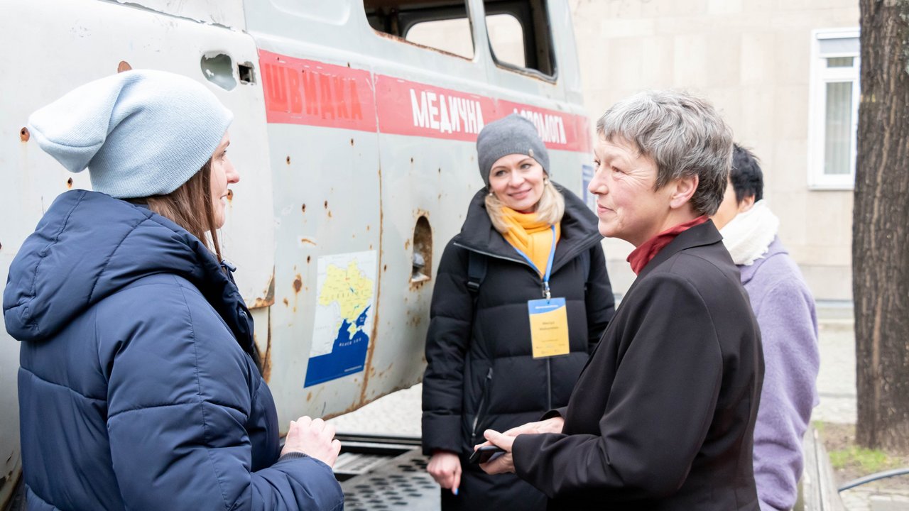Hanna Naber im Gespräch mit zwei Vertreterinnen der Organisation Fellas for Europe e.V. neben dem schwer beschädigten Krankenwagen