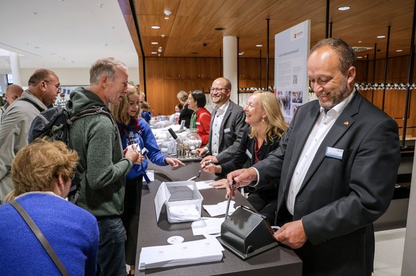 Bild: © Focke Strangmann Links einige Besucherinnen und Besucher, ihnen gegenüber drei lachende Mitarbeitende des Stenografischen Dienstes mit Button-Maschine, Notizblock und Stanze.