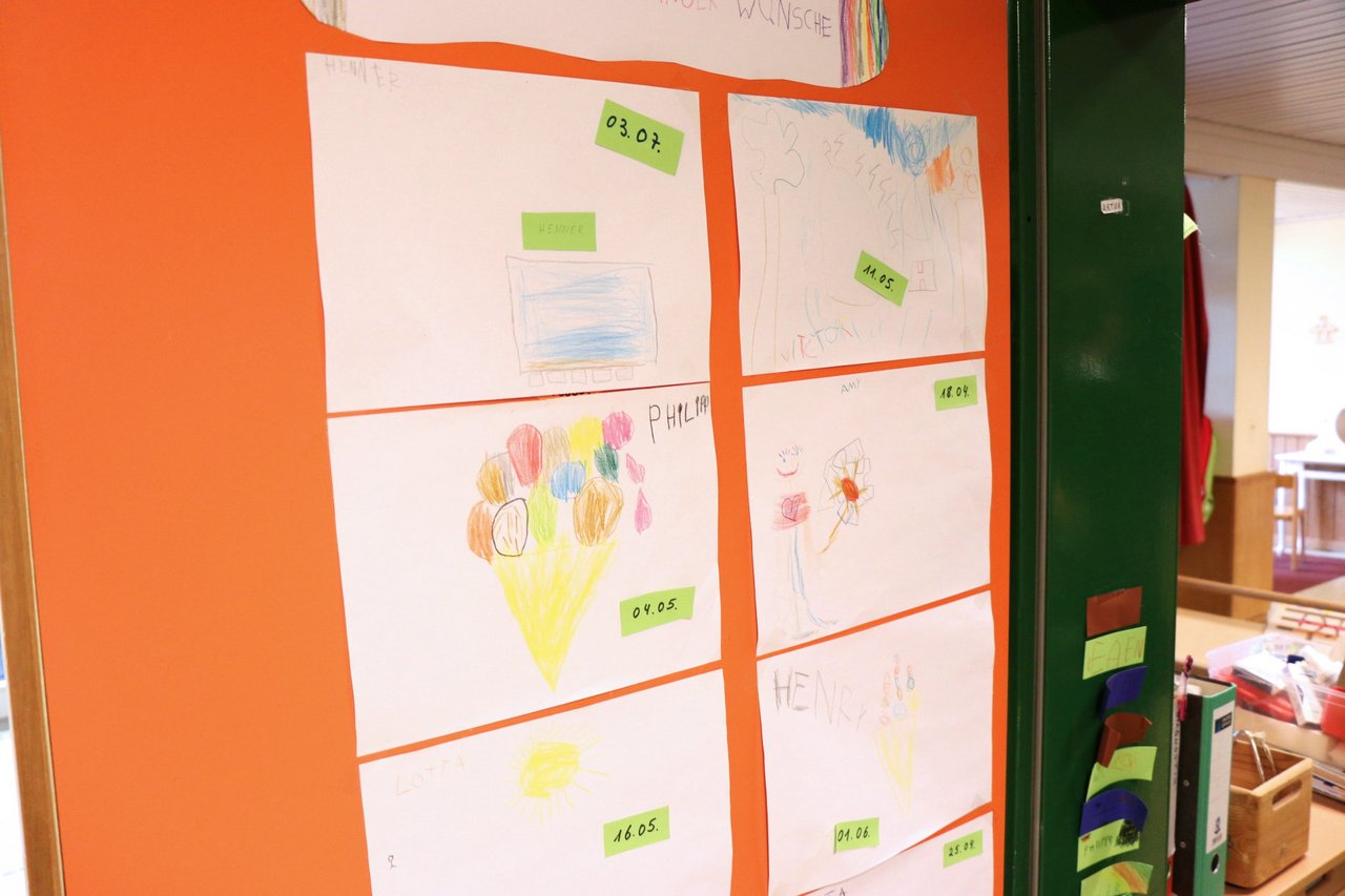 Von Kindern gemalte Bilder an einer orangefarbenen Pinnwand.
