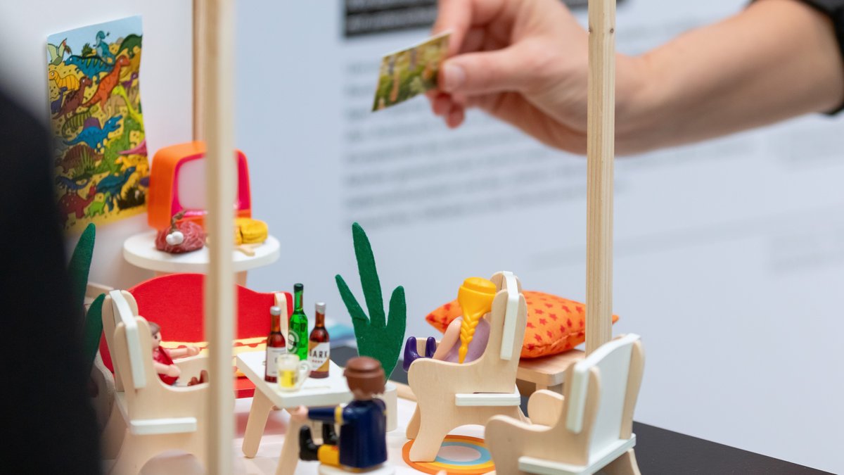 Nahaufnahme einer Hand, die Objekte in einem Spielzeugmodell eines Wohnzimmers umstellt.