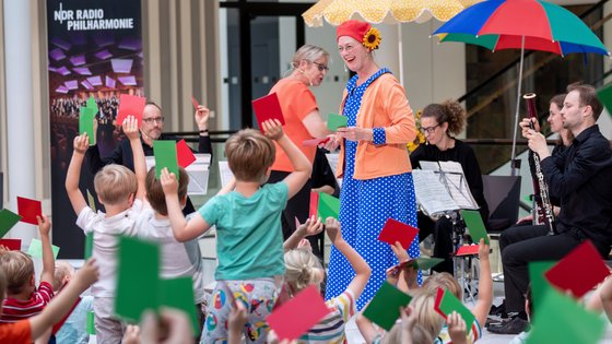 Foto aus der Portikushalle. Im Vordergrund sind viele Kinder zu sehen, die mit dem Rücken zur Kamera auf bunten Kissen auf dem Boden sitzen. Ihnen gegenüber stehen Susanne Grünig sowie die Musikerinnen und Musiker der NDR Radiophilharmonie mit Instrumenten und Notenständern.