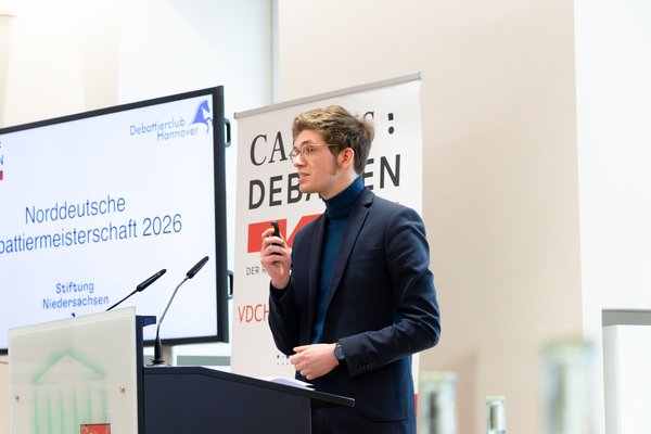 Ein junger Mann im Sakko steht am Redepult. Im Hintergrund ein Bildschirm mit der Aufschrift: Norddeutsche Debattiermeisterschaft 2026.