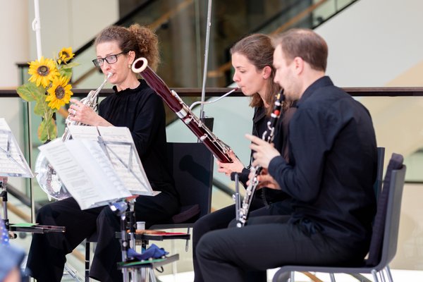 Zwei Musikerinnen und ein Musiker sitzen in der Portikushalle, schauen auf ihre Notenblätter und spielen Musik auf ihren Blasinstrumenten. Im Hintergrund sind ein Teil der Niedersachsentreppe und Blumendekoration zu erkennen.