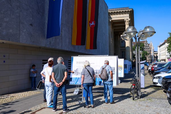 Klick auf das Bild öffnet eine vergrößerte Ansicht in einem Overlay. Zum Schließen des Overlays die ESC-Taste drücken. Eine Ausstellung aus großen Planen vor dem Landtagsgebäude mit mehreren Menschen, die sie sich anschauen.'}