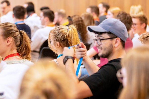 Bild: © Niedersächsischer Landtag Publikum bestehend aus jungen Menschen, die bunte Schlüsselbänder tragen, im Vordergrund hängt sich ein junger Mann ein blaues Band um.