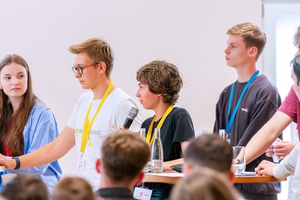 Klick auf das Bild öffnet eine vergrößerte Ansicht in einem Overlay. Zum Schließen des Overlays die ESC-Taste drücken. Vier Jugendliche währen der Abschlussdiskussion, eine von ihnen spricht ins Mikrofon.'}