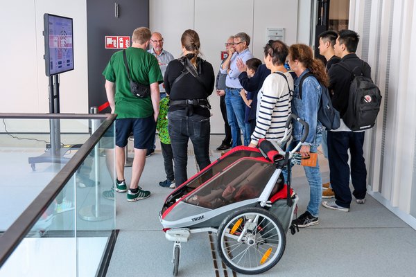 Klick auf das Bild öffnet eine vergrößerte Ansicht in einem Overlay. Zum Schließen des Overlays die ESC-Taste drücken. Eine Gruppe von Besucherinnen und Besuchern an einer Info-Station, im Vordergrund steht ein Kinderwagen.'}