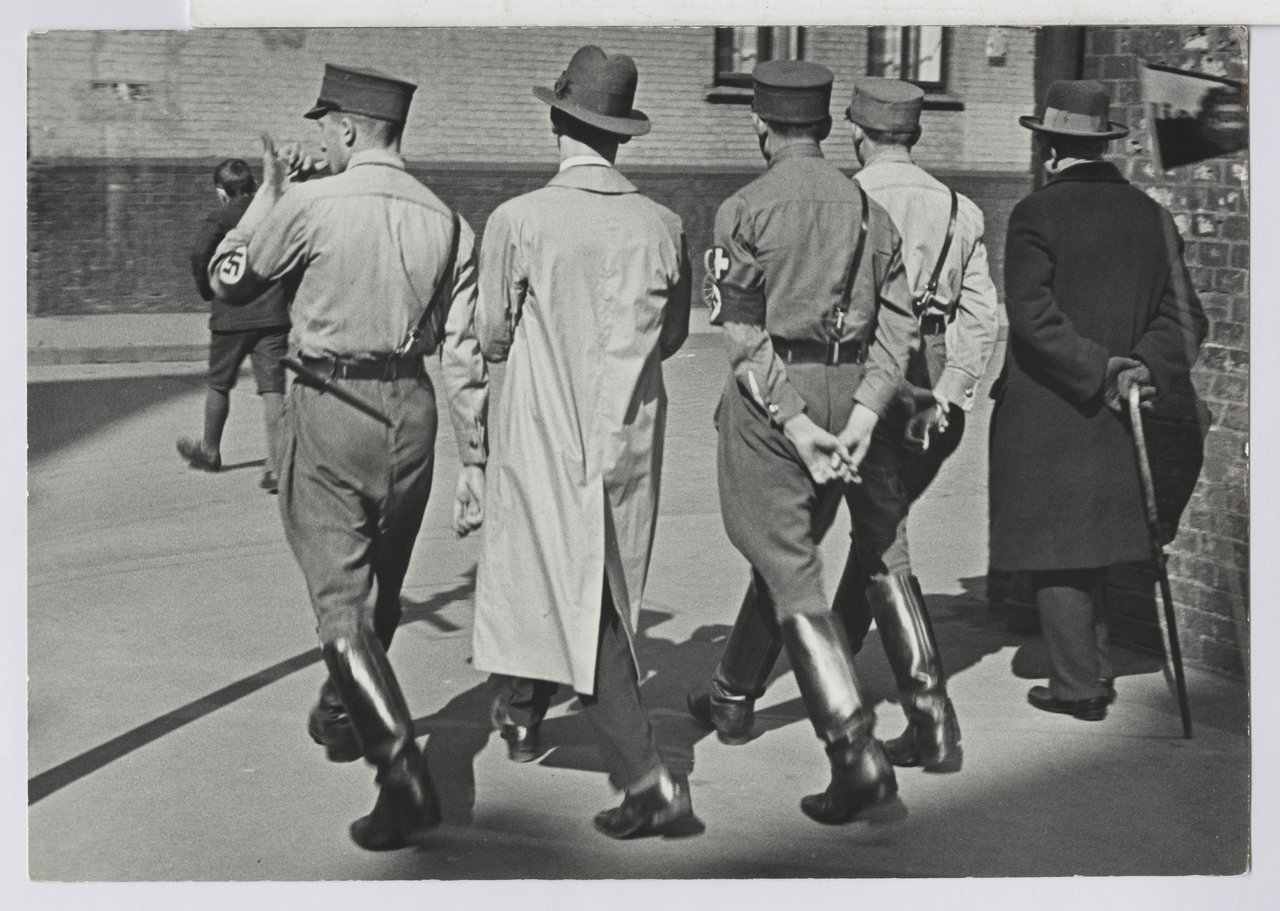 Fünf Männer mit NS-Uniformen von hinten im Gleichschritt.