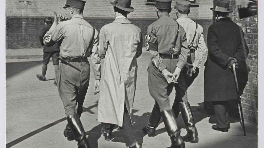 Fünf Männer mit NS-Uniformen von hinten im Gleichschritt.