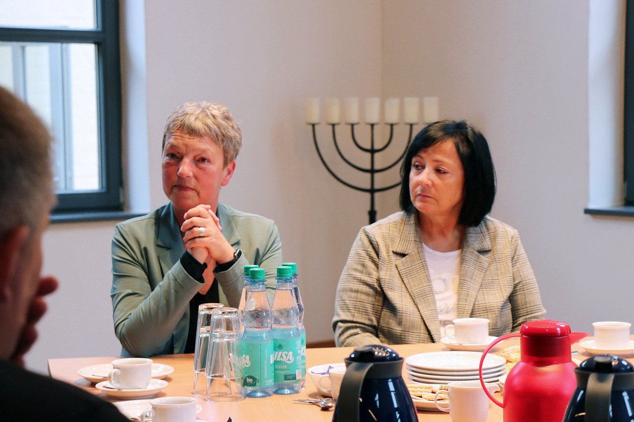 Landtagspräsidentin Naber links neben einer Frau am Tisch, sie hören jemandem außerhalb des Bildes zu; im Hintergrund eine Menora.