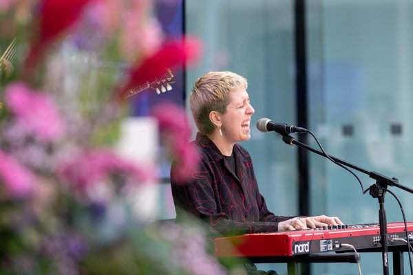 Bild: © Niedersächsischer Landtag Die Sängerin der Band Hertzcasper spielt E-Piano und singt ins Mikrofon. Im Vordergrund sind einige Blüten eines Blumenstraußes in Lila- und Pinktönen zu sehen.