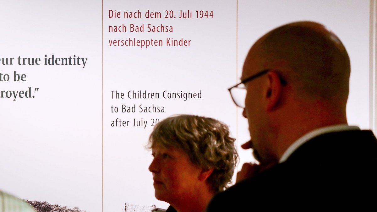 Landtagspräsidentin Hanna Naber und ein Mann in der Ausstellung. Im Hintergrund ist eine weiße Fläche mit einem Schwarz-Weiß-Foto und Text in Deutsch und Englisch zu sehen. Der deutsche Text lautet: "Die nach dem 20. Juli 1944 nach Bad Sachsa verschleppten Kinder"
