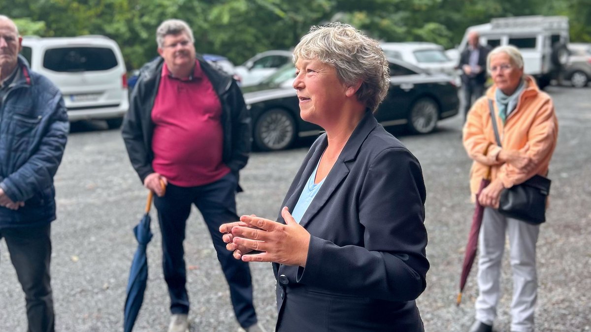 Landtagspräsidentin Hanna Naber steht vor Beginn der Wanderung auf einem Parkplatz und spricht zu den Anwesenden. Im Hintergrund sind mehrere Menschen zu sehen.