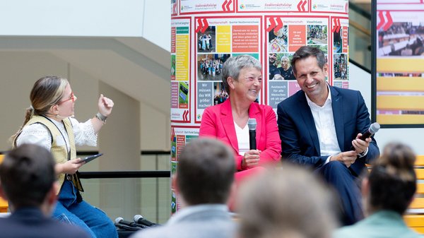 Bild: © Niedersächsischer Landtag Landtagspräsidentin Hanna Naber und Minister Olaf Lies sitzen lachend neben der Moderatorin auf der Bühne. Im Vordergrund sieht man unscharf ein paar Hinterköpfe des Publikums.