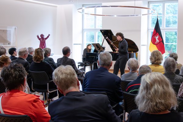 Bild: © Niedersächsischer Landtag Ein junger Musiker spielt ein Fagott vor einer Gruppe von Zuhörern in einem hellen Raum mit großen Fenstern und einem Konzertflügel, auf dem eine junge Musikerin spielt. Im Vordergrund ist ein Teil des Publikums von hinten zu sehen.