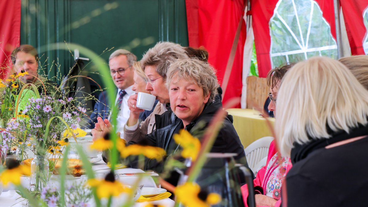 Landtagspräsidentin Hanna Naber sitzt mit vielen anderen Menschen an einer langen Tafel mit Blumensträußen, Kaffeetassen und Kuchen.