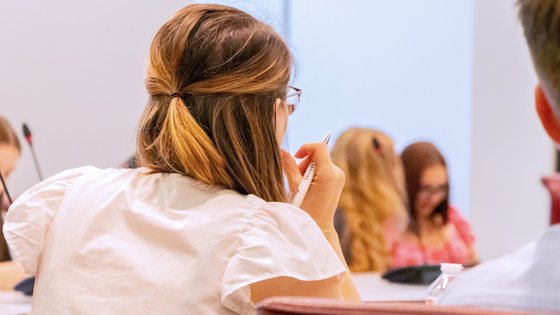 Eine junge Frau mit einem Stift in der Hand, an einem Tisch sitzend, von hinten, im Hintergrund weitere Personen.