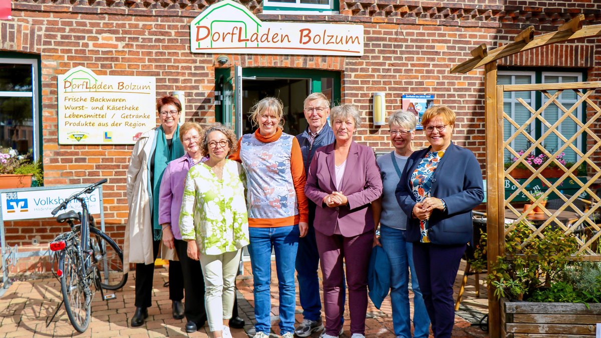 Gruppenfoto von Landtagspräsidentin Hanna Naber und dem Team des Dorfladen Bolzum. Die Gruppe steht draußen vor dem Dorfladen in der Sonne, im Hintergrund sind die Backsteinfassade und Schilder des Ladens zu sehen.
