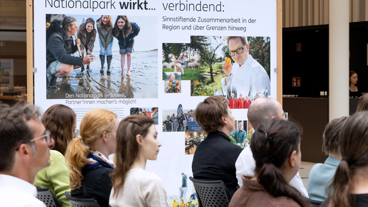 Eine Personengruppe sitzt vor einem Aufsteller über den Nationalpark mit Fotos von Naturerlebnissen und der Überschrift "Nationalpark wirkt verbindend".
