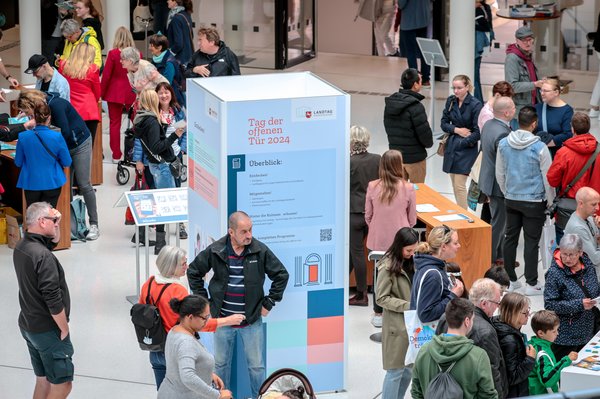 Bild: © Focke Strangmann Ansicht der gut besuchten Portikushalle beim Tag der offenen Tür. In der Mitte steht eine Säule mit dem Text "Tag der offenen Tür 2024" sowie Informationen zum Programm.