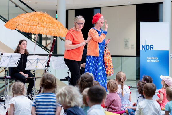 Susanne Grünig und Frau Muse (alias Heidi Vollprecht) stehen vor den Kindern und winken ihnen fröhlich zu. Links im Hintergrund sitzt eine der Musikerinnen der NDR Radiophilharmonie, rechts steht ein Roll-Up des NDR.