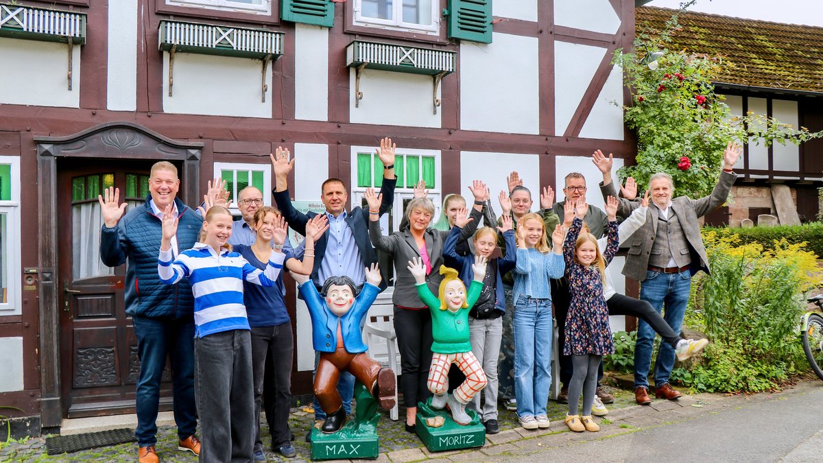 Landtagspräsidentin Hanna Naber steht mit einer größeren Gruppe Menschen vor der Wilhelm-Busch-Mühle. In vorderster Reihe stehen zwei Statuen von Max und Moritz, die die Arme nach oben recken. Die Gruppe tut es ihnen gleich.