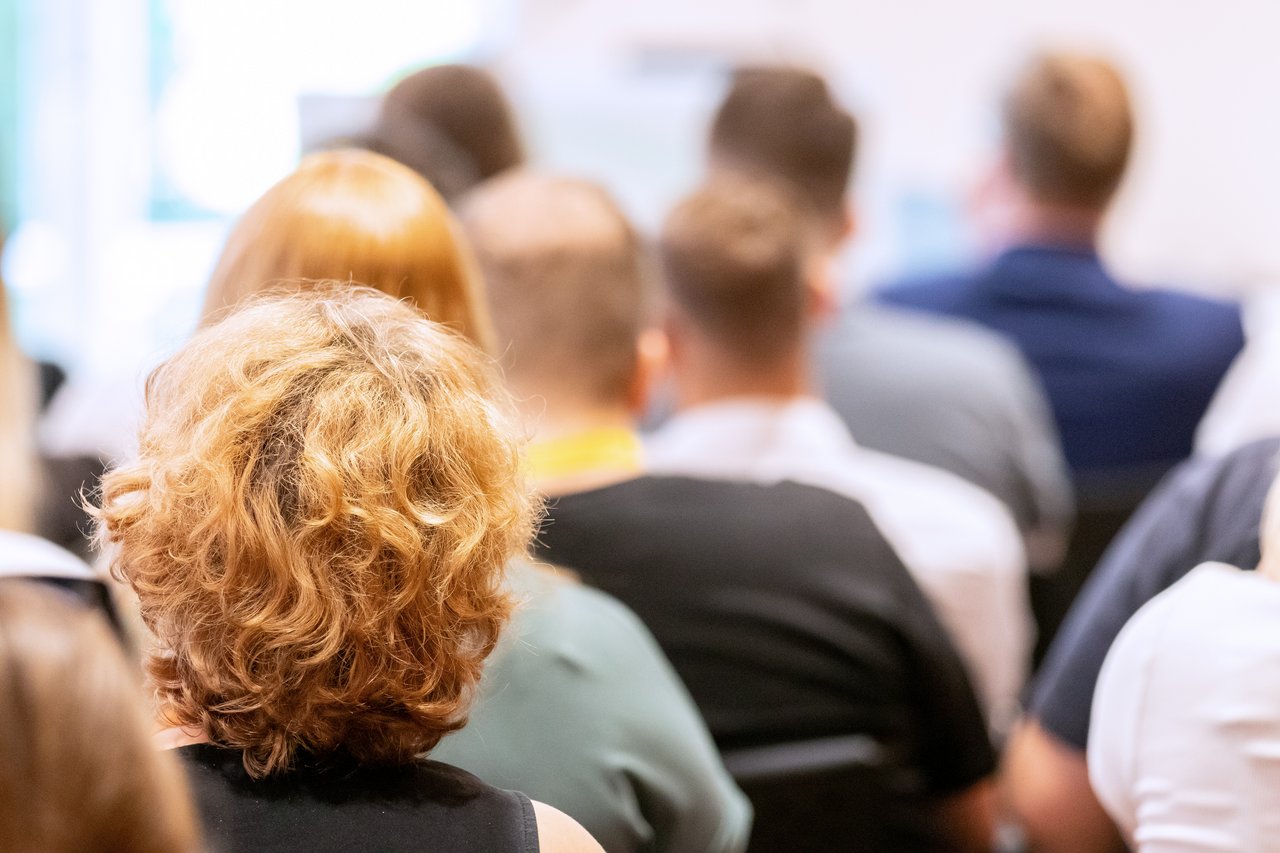 Bild: © Niedersächsischer Landtag Viele Personen sitzend von hinten; eine Frau mit lockigem Haar im Fokus.