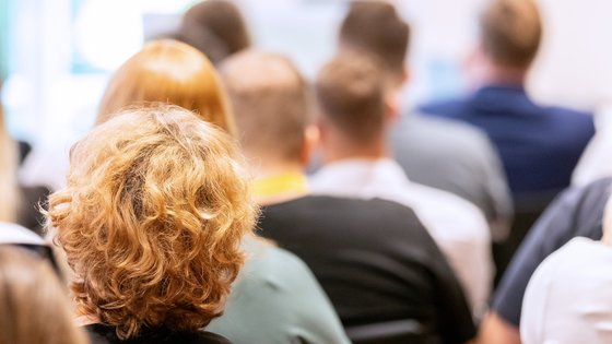 Viele Personen sitzend von hinten; eine Frau mit lockigem Haar im Fokus.