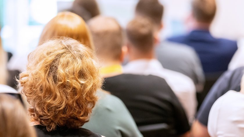 Viele Personen sitzend von hinten; eine Frau mit lockigem Haar im Fokus.