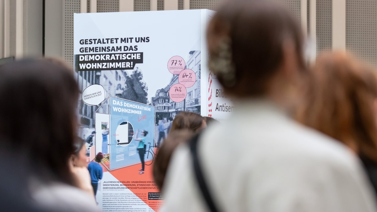 Im Vordergrund unscharf das Publikum, im Hintergrund fokussiert das Plakat mit dem Titel „Das demokratische Wohnzimmer“