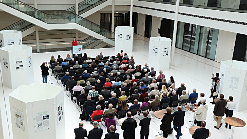 Voll besetzte Sitzreihen in der Portikushalle. Die Gäste der Veranstaltung nehmen inmitten der Ausstellung Platz.