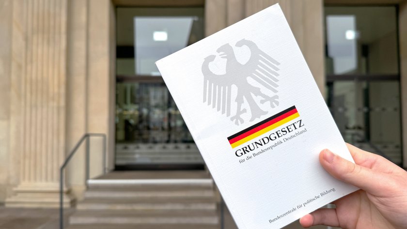 Grundgesetz, das in einer Hand hochgehalten wird. Dahinter der Portikus des Niedersächsischen Landtages.