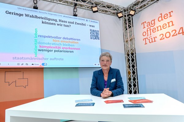Bild: © Focke Strangmann Landtagspräsidentin Hanna Naber steht an einem Stehtisch und lächelt in die Kamera. Im Hintergrund ist links das Ideenboard mit der Aufschrift "Geringe Wahlbeteiligung, Hass und Zweifel - was können wir tun?" zu sehen und rechts der Schriftzug "Tag der offenen Tür 2024".