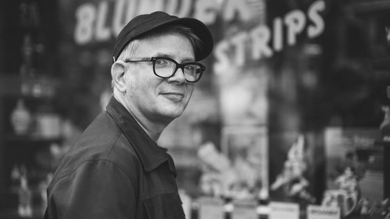 Portraitfoto von Tobi Dahmen in Schwarz-Weiß. Er hat helle Haare, trägt eine Brille mit dunklem Gestell, eine dunkle Kappe und eine dunkle Jacke. Er steht seitlich zur Kamera und dreht den Kopf mit einem leichten Lächeln zum Betrachter. Im Hintergrund ist eine große Schaufensterfront zu erkennen.
