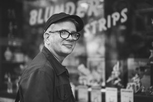 Portraitfoto von Tobi Dahmen in Schwarz-Weiß. Er hat helle Haare, trägt eine Brille mit dunklem Gestell, eine dunkle Kappe und eine dunkle Jacke. Er steht seitlich zur Kamera und dreht den Kopf mit einem leichten Lächeln zum Betrachter. Im Hintergrund ist eine große Schaufensterfront zu erkennen.