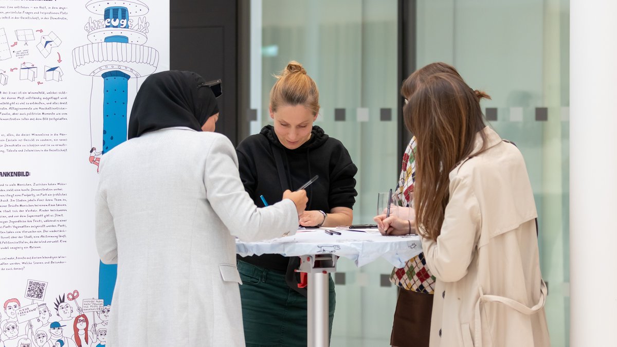 Studierende und Gäste stehen an einem Tisch vor einer Ausstellungswand und zeichnen.