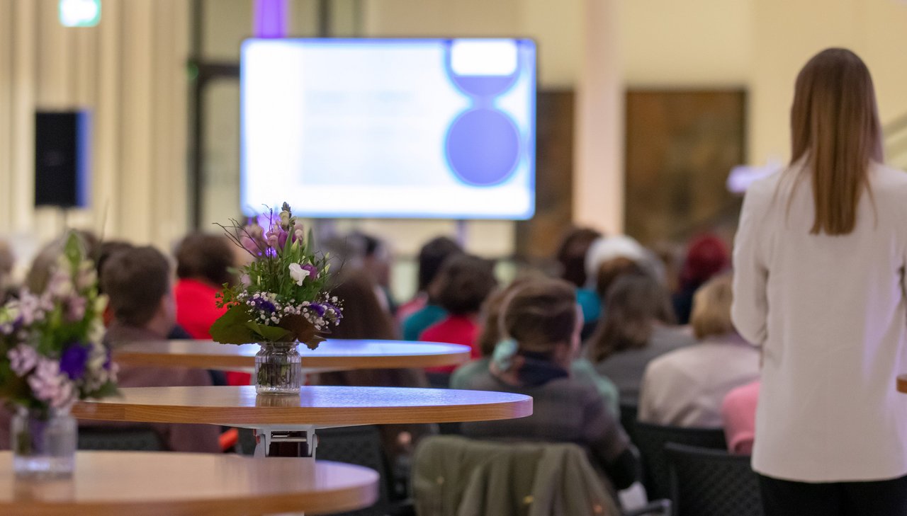 Im Vordergrund mit Blumendekoration geschmückte Stehtische, dahinter das Publikum