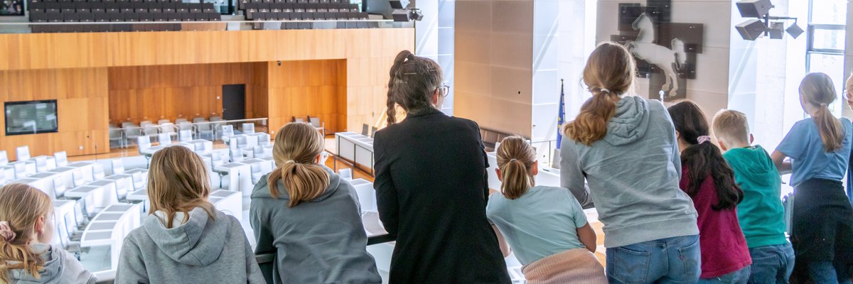 Eine Landtagslotsin umringt von Kindern schaut mit ihnen von der Besuchertribüne hinunter in den leeren Plenarsaal. 