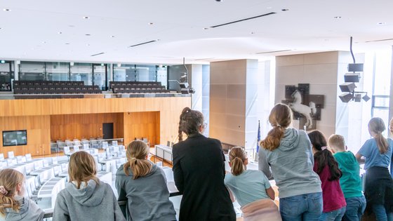 Eine Landtagslotsin umringt von Kindern schaut mit ihnen von der Besuchertribüne hinunter in den leeren Plenarsaal. 