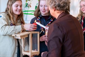 Eine Person in Pfadfinderuniform hält eine Kerze, Landtagspräsidentin Naber entzündet sie. Im Hintergrund eine weitere Person in Pfadfinderuniform.