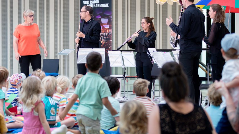 Foto aus der Portikushalle. Im Vordergrund sind viele Kinder zu sehen, die mit dem Rücken zur Kamera auf bunten Kissen auf dem Boden sitzen. Ihnen gegenüber stehen Susanne Grünig sowie die Musikerinnen und Musiker der NDR Radiophilharmonie mit Instrumenten und Notenständern.