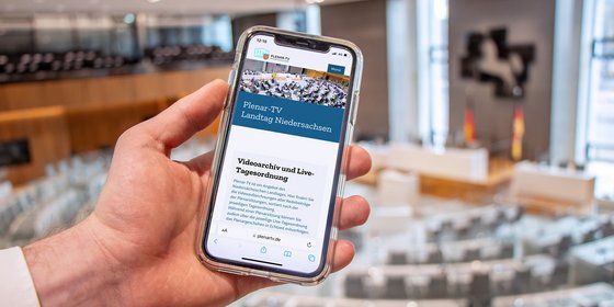 Auf einem Smartphone, das in der Hand einer Person liegt, ist die Seite "Plenar-TV" geöffnet.