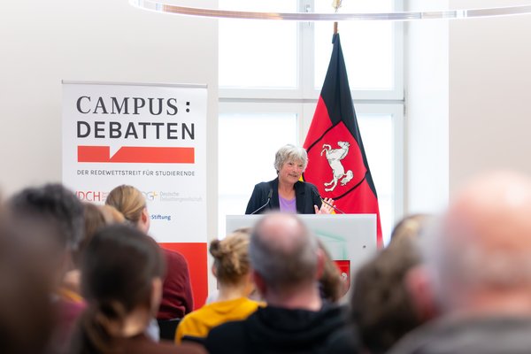 Landtagspräsidentin Hanna Naber am Redepult im Leibniz-Saal. Neben ihr steht ein Aufsteller mit dem Logo der Campus-Debatten, im Vordergrund ist ein Teil des Publikums von hinten zu erkennen.