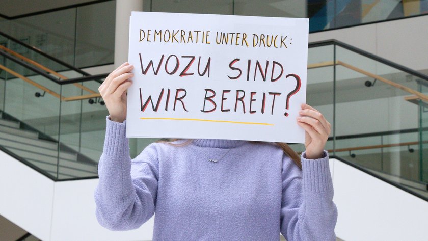 Person hält ein Schild hoch mit dem handgeschriebenen Schriftzug: Demokratie unter Druck. Wozu sind wir bereit?