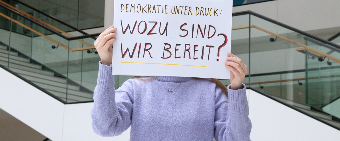Person hält ein Schild hoch mit dem handgeschriebenen Schriftzug: Demokratie unter Druck. Wozu sind wir bereit?