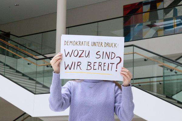 Person hält ein Schild hoch mit dem handgeschriebenen Schriftzug: Demokratie unter Druck. Wozu sind wir bereit?