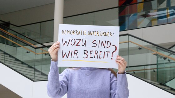 Person hält ein Schild hoch mit dem handgeschriebenen Schriftzug: Demokratie unter Druck. Wozu sind wir bereit?
