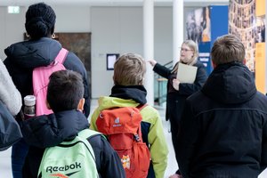 Schülerinnen und Schüler stehen mit dem Rücken zur Kamera in der Portikushalle. Im Hintergrund steht eine junge Frau und spricht zu ihnen.