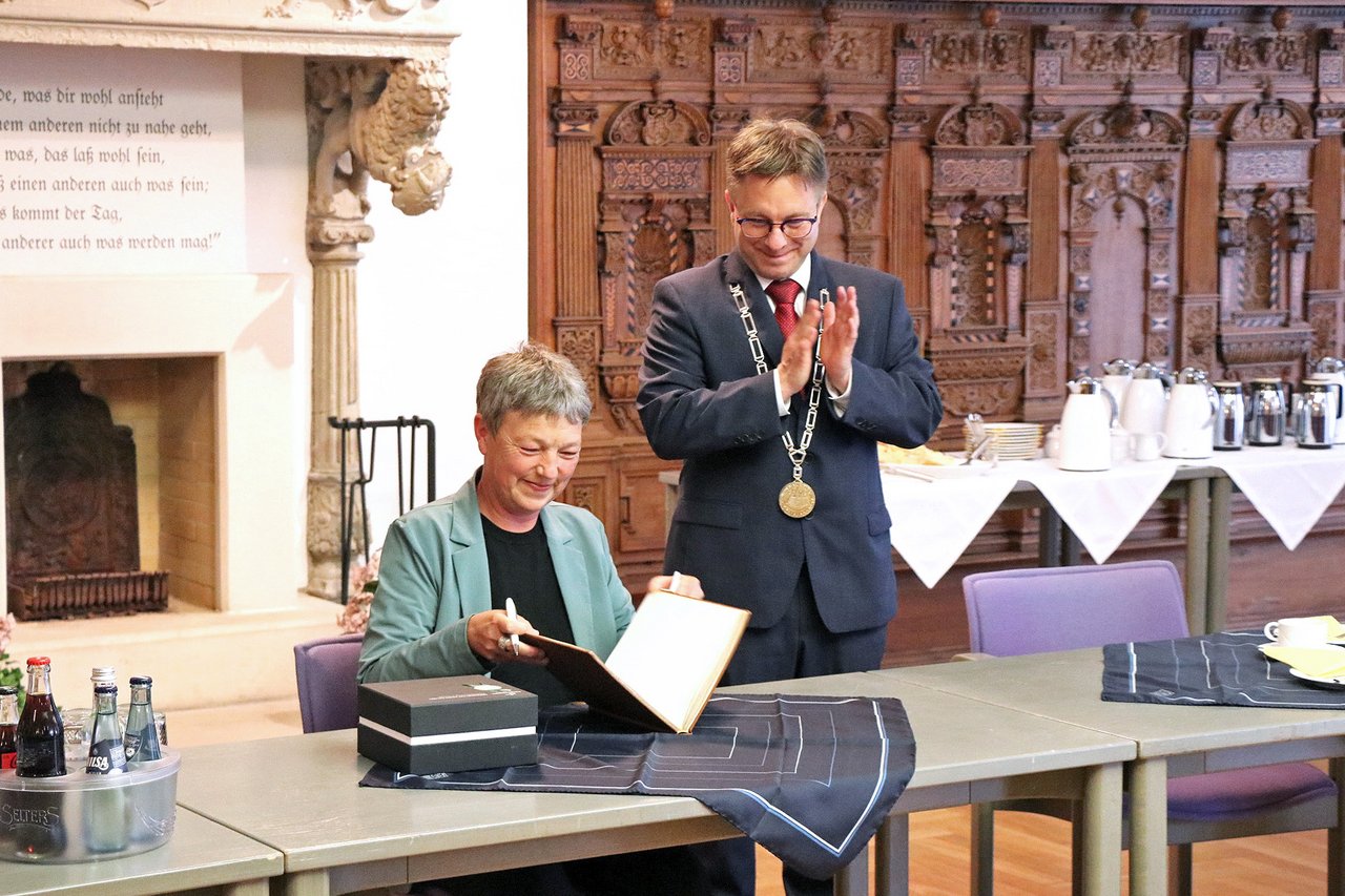 Bild: © Niedersächsischer Landtag Landtagspräsidentin Naber im Rathaus von Jever, sie schlägt gerade das goldene Buch der Stadt zu. Neben ihr Bürgermeister Jan Edo Albers, klatschend.