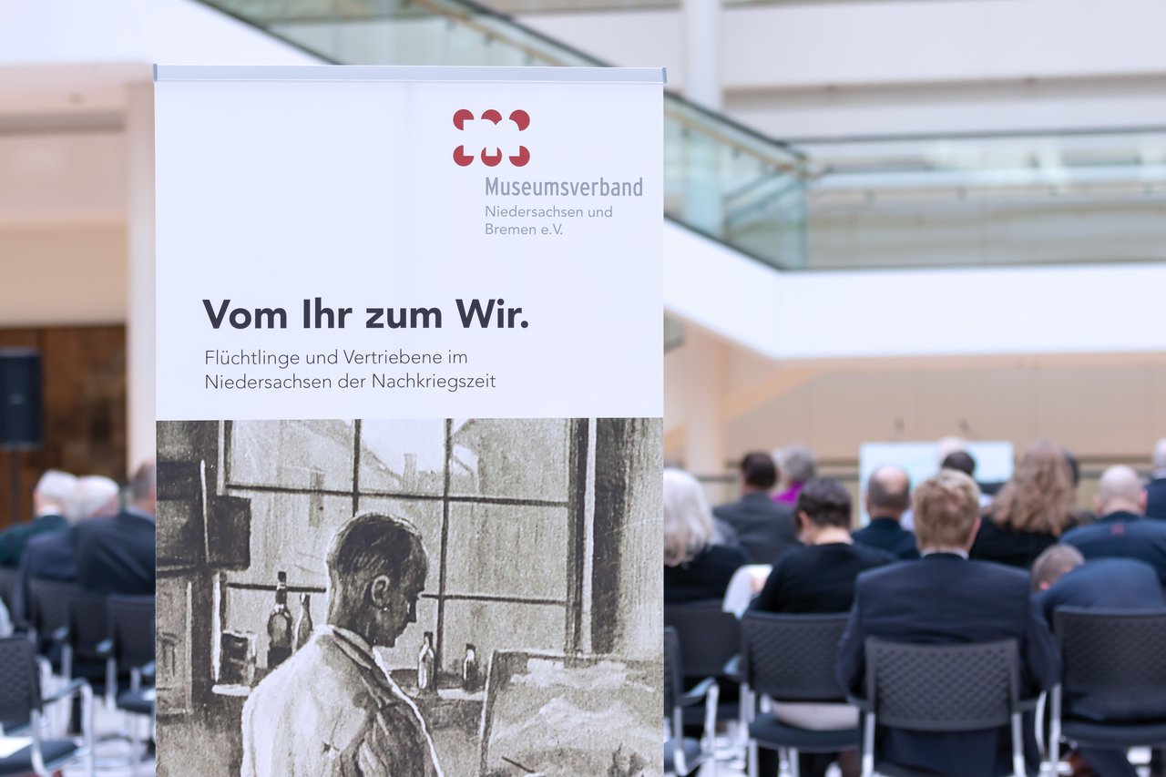 Banner mit dem Titel der Ausstellung, im Hintergrund das Publikum der Ausstellungseröffnung